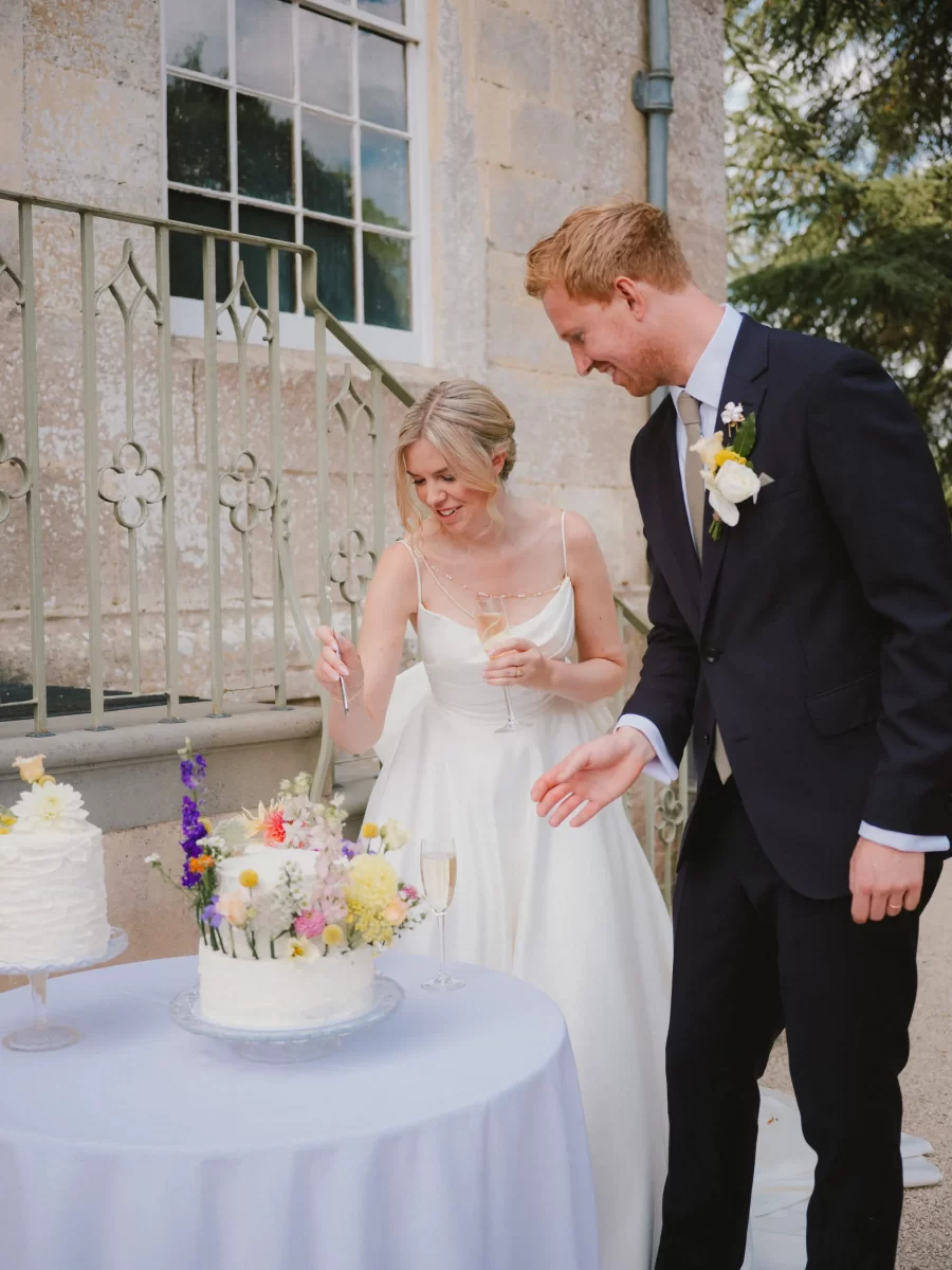 A bride and groom stand by a table with decorated cakes outdoors. The bride, holding a knife and a champagne glass, prepares to cut the main cake while the groom, in a suit, smiles beside her.