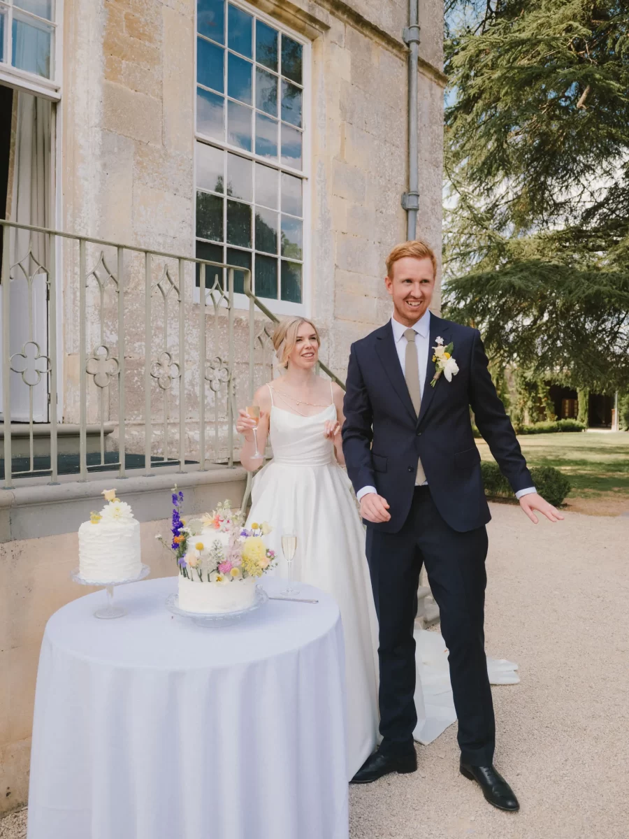 A bride in a white dress and a groom in a navy suit stand smiling outdoors by a table with two small wedding cakes decorated with flowers, near a stone building with large windows and greenery in the background.