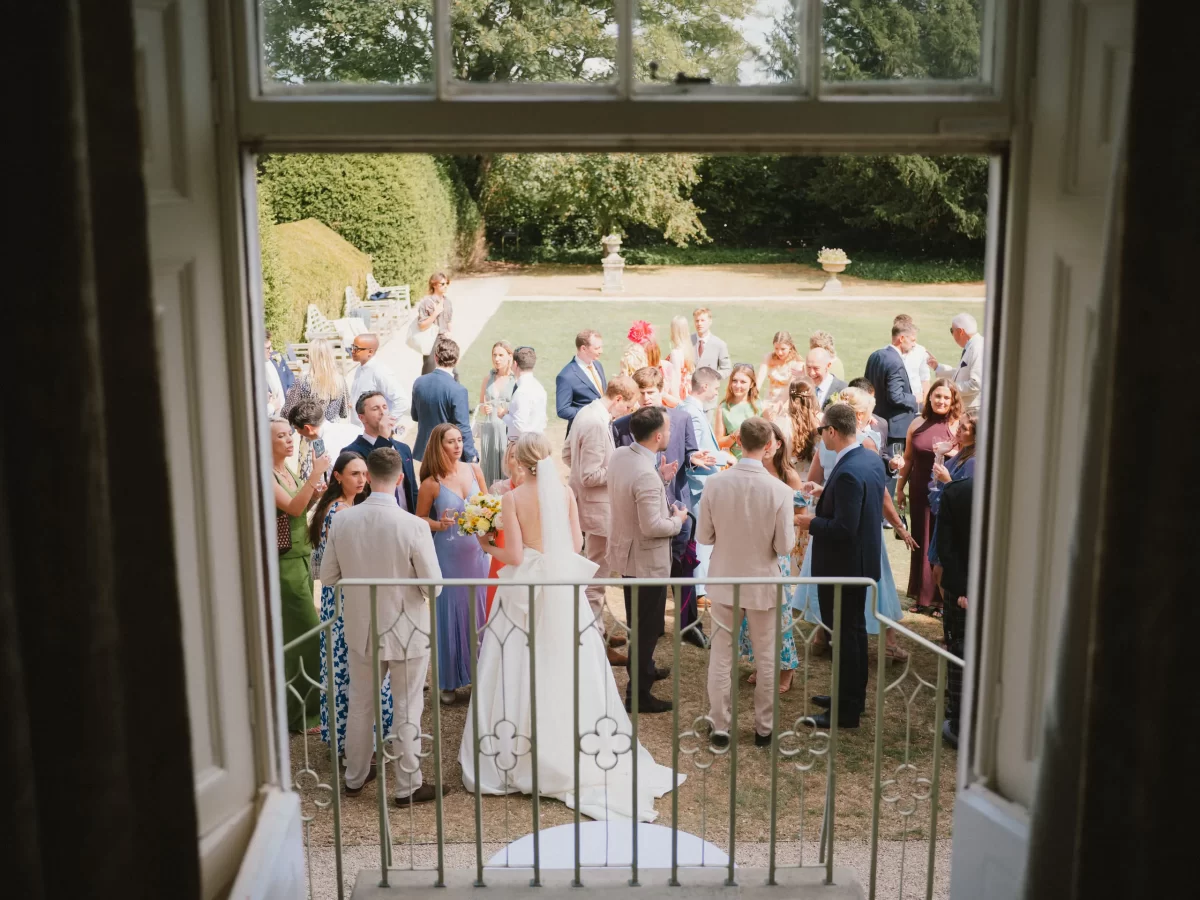 View from a window overlooking a garden at Elmore court where a group of people, including a bride in a white dress, are gathered and socializing at an outdoor wedding reception on a sunny day.
