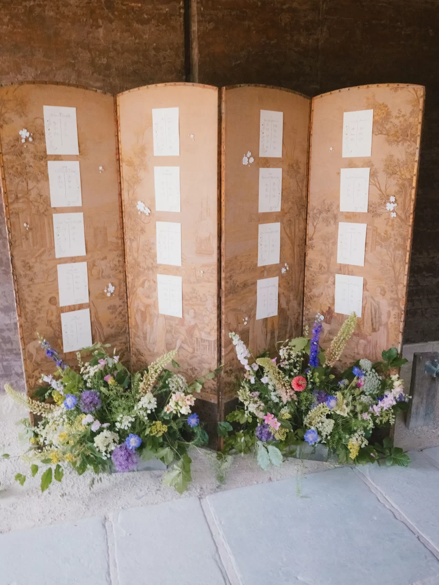 A vintage folding screen decorated with papers and small white flower cutouts stands against a wall. Colorful wildflower arrangements are placed at the base of the screen on a stone floor.