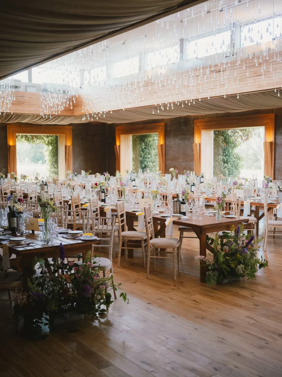 A spacious, elegantly decorated dining hall with wooden floors, many tables set with glasses and plates, wooden chairs, floral centerpieces, and large windows letting in natural light. Gillyflower at Elmore Court
