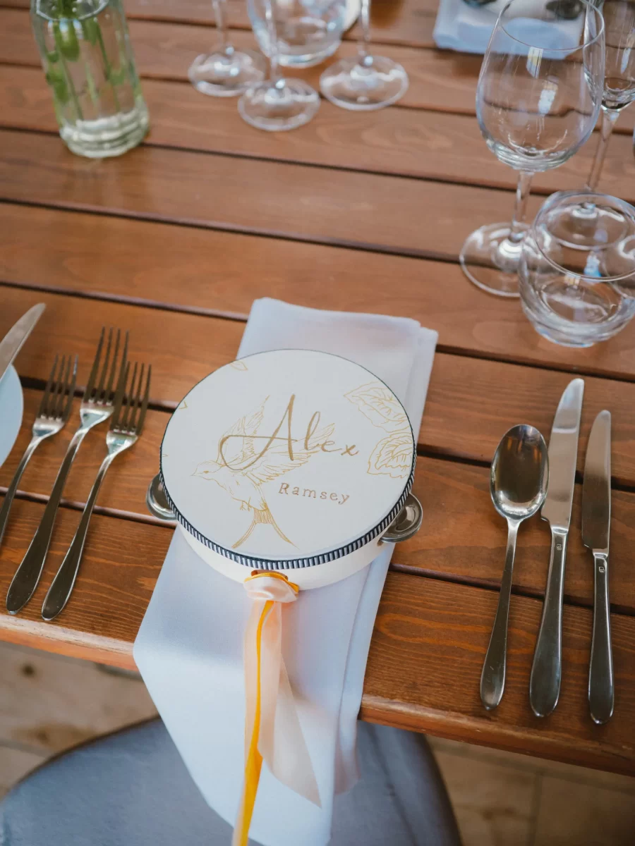 A round box with the name Alex Ramsey in gold lettering sits on a white napkin at a wooden table set with cutlery, glasses, and a ribbon, likely for a formal event or celebration.