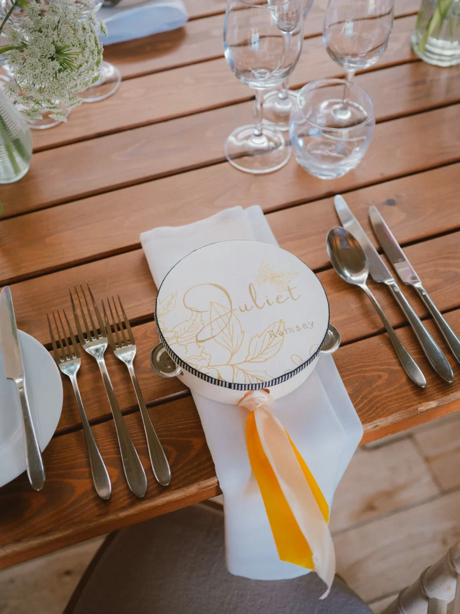 A decorated place setting at a wooden table, featuring a menu or name card with Juliet written on it, yellow and white ribbons, silver cutlery, and glassware arranged neatly.