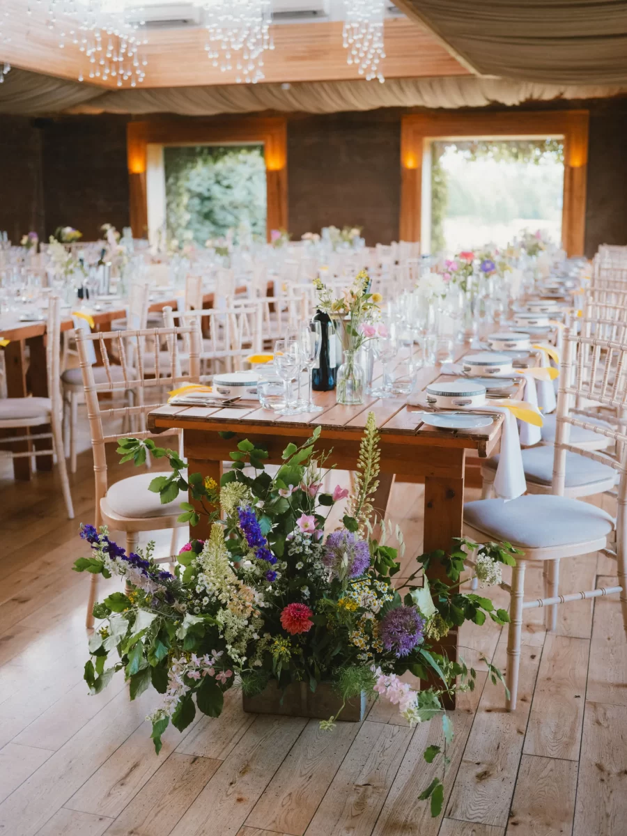 A long wooden table set for a formal event with plates, glasses, and napkins, surrounded by white chairs and decorated with wildflower bouquets; large floral arrangement at the table’s end in a bright, airy room.