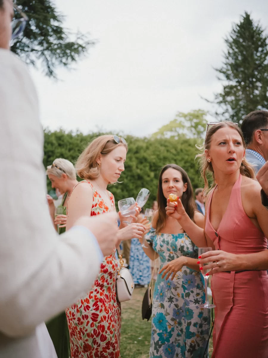 A group of people dressed in summer outfits stand outdoors, holding drinks and talking. A woman in a pink dress gestures while others listen, with greenery and trees in the background.
