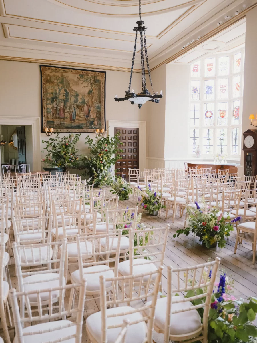 A bright, elegant room set up for a wedding ceremony with rows of white chairs, floral arrangements lining the aisle, tall windows letting in natural light, and a tapestry and chandelier overhead.