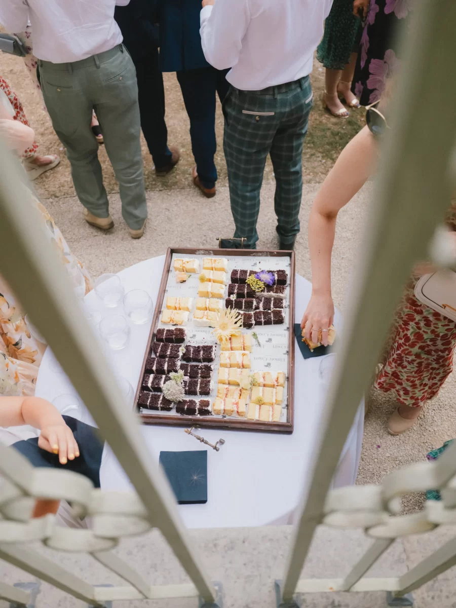 A tray of assorted desserts sits on a round table outdoors, surrounded by people dressed in semi-formal attire. The scene is viewed from above, framed by white railings in the foreground.