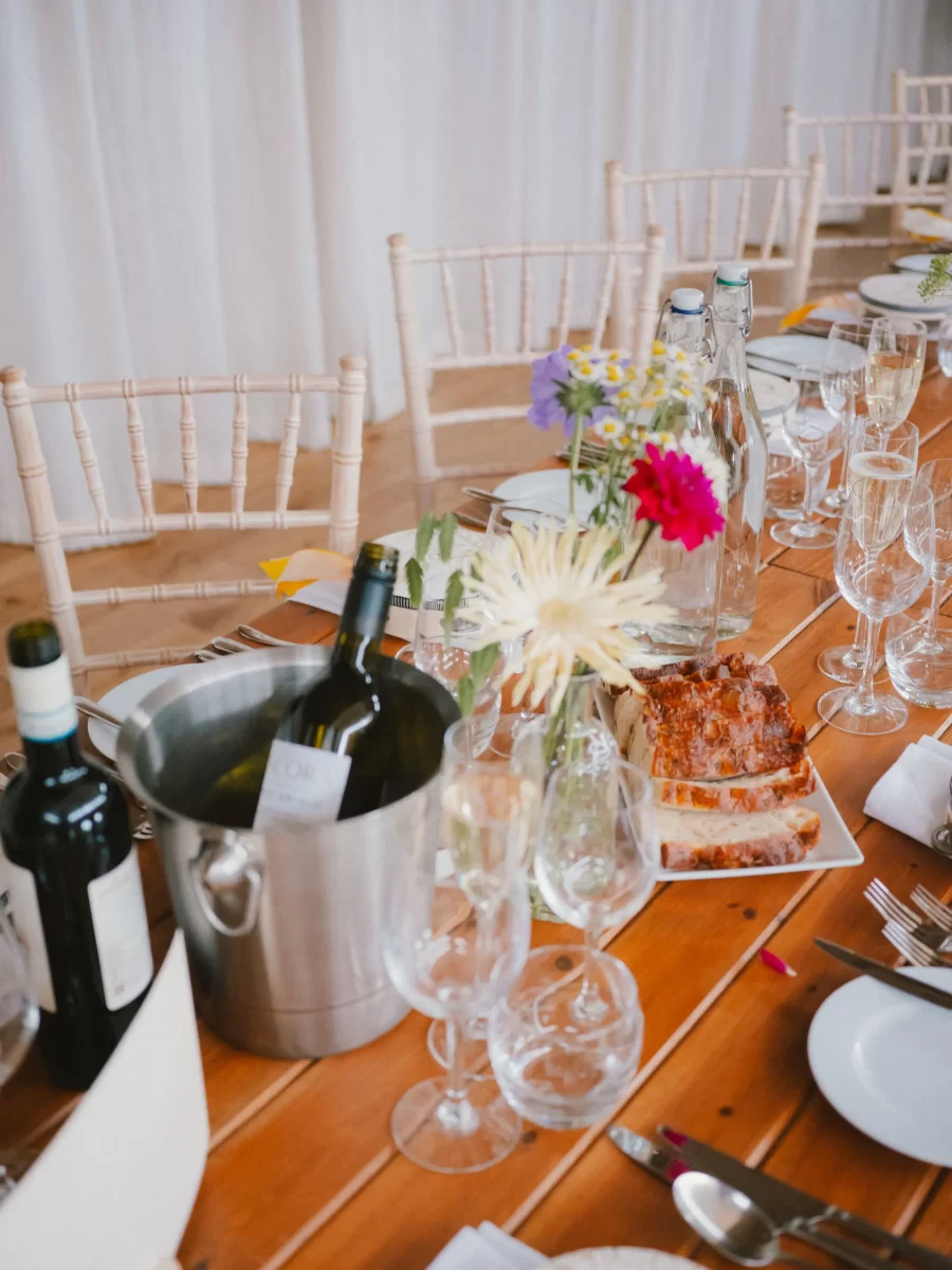 A rustic wooden table set for a meal with wine bottles, glasses, plates, a metal ice bucket, water bottles, a vase of colorful flowers, and a loaf of bread on a platter. Pale chairs and white drapes are in the background.