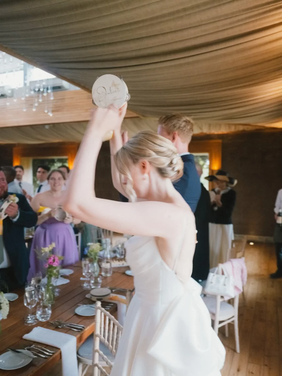 A bride in a white dress raises a plate above her head, surrounded by guests at a decorated indoor wedding reception with wooden floors and draped ceilings.