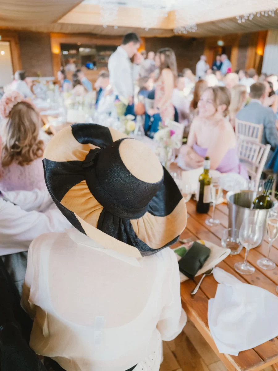 A person wearing a large black and beige hat sits at a crowded, festive table during a lively indoor event, surrounded by people dressed in light, elegant clothing.