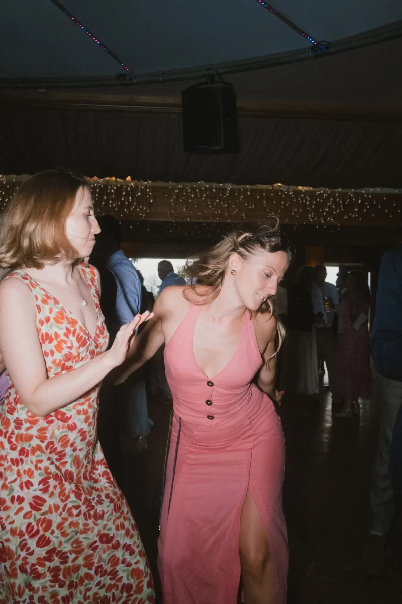 Two women in colorful dresses dancing together at an indoor event with string lights in the background. One wears a floral dress, the other a pink dress with buttons and a slit. Other people are visible in the dimly lit background.