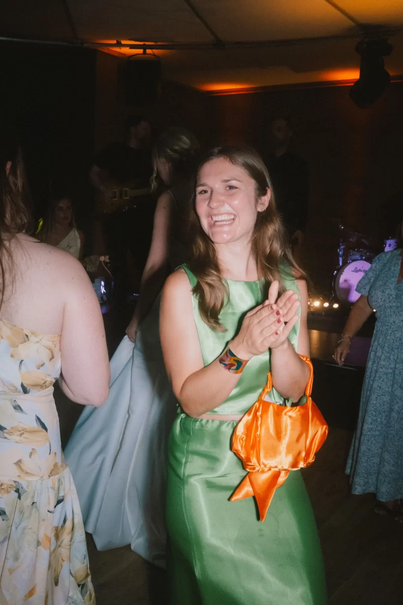 A woman in a shiny green dress smiles and claps while holding an orange bag at a lively indoor event, surrounded by other people dancing under warm lights.