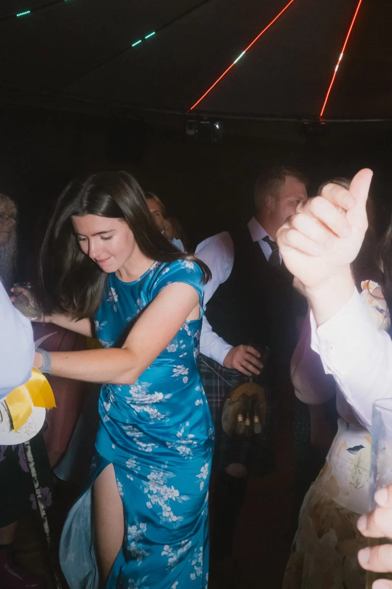 A woman in a blue floral dress dances energetically at a lively indoor event, surrounded by other people. Colorful lights create a festive atmosphere, and a raised hand with a thumbs-up is visible in the foreground.