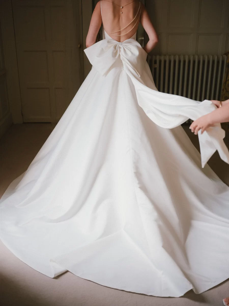 A woman in a white, backless wedding gown with a large bow on the back stands indoors; another person’s hand is gently holding part of the dress’s train.