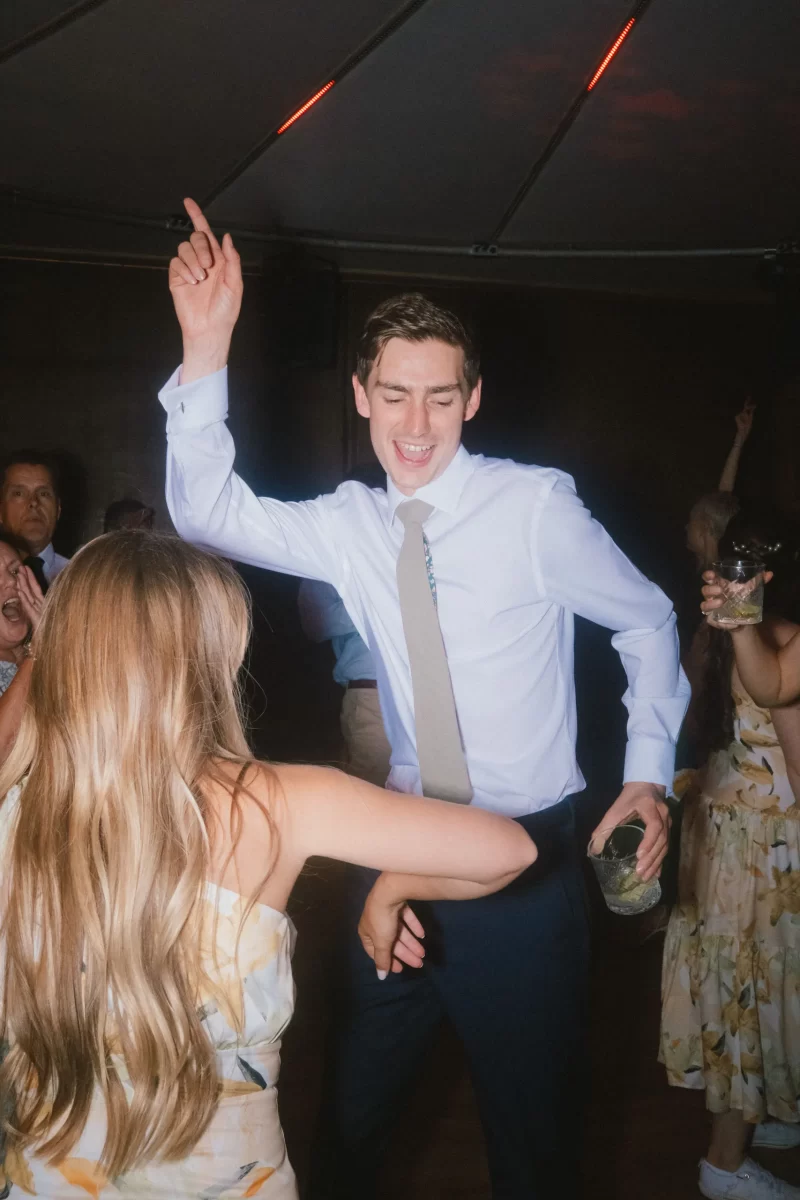 A young man in a white shirt and tie dances enthusiastically with a drink in hand at a lively indoor party, surrounded by people in floral attire.