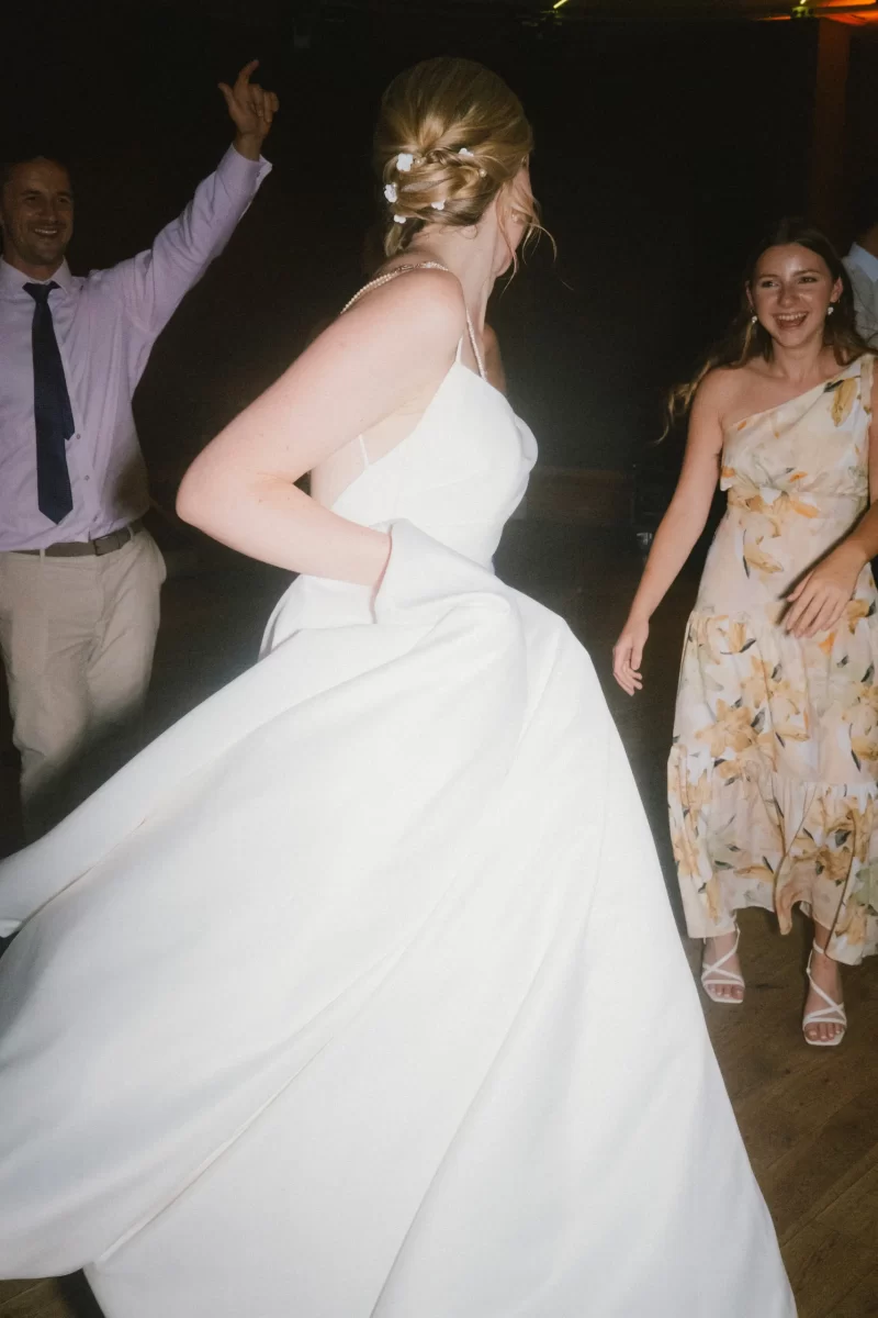 A bride in a white gown twirls on a dance floor, her back turned to the camera. A smiling man and a woman in a floral dress stand nearby, enjoying the celebration.