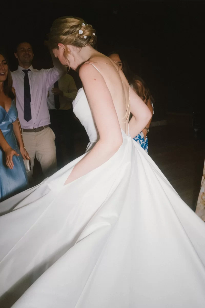 A bride in a white gown with a low back and floral hairpiece spins on the dance floor, surrounded by smiling guests in formal attire at Elmore Court