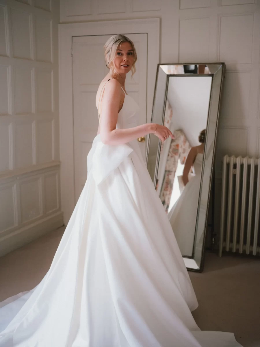 A woman in a white wedding dress stands indoors, looking over her shoulder. She is in front of a large mirror, and the room has paneled walls and a radiator. Natural light fills the space.