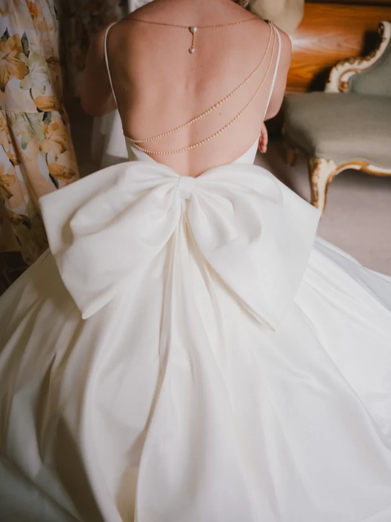 A bride is shown from behind, wearing a white wedding dress with a large bow at the back and delicate gold chain jewelry draped across her bare back. She is seated indoors near vintage furniture at Elmore Court