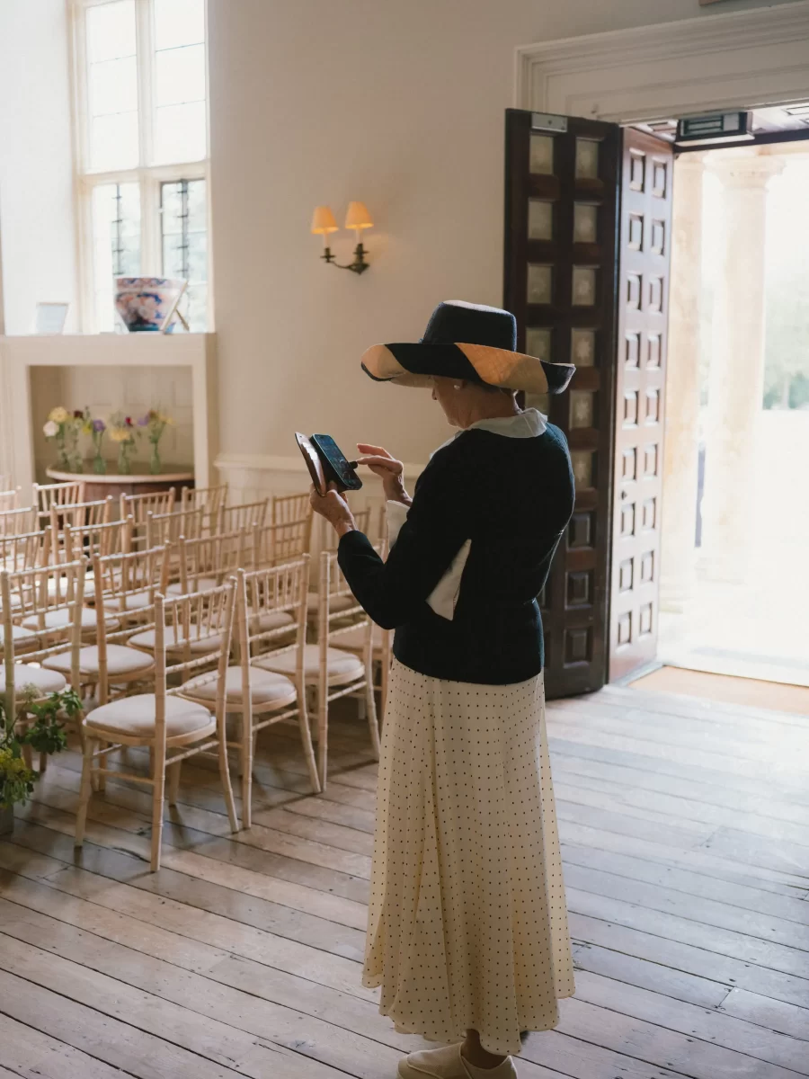 A woman in a wide-brimmed hat and long skirt stands indoors near empty wooden chairs, using a tablet or phone. Sunlight streams through an open door beside her, creating a bright, airy atmosphere.