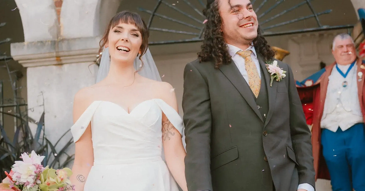 A joyful bride in a white dress and a smiling groom in a dark suit stand hand in hand outside, celebrating their George in Rye wedding with confetti falling around them.