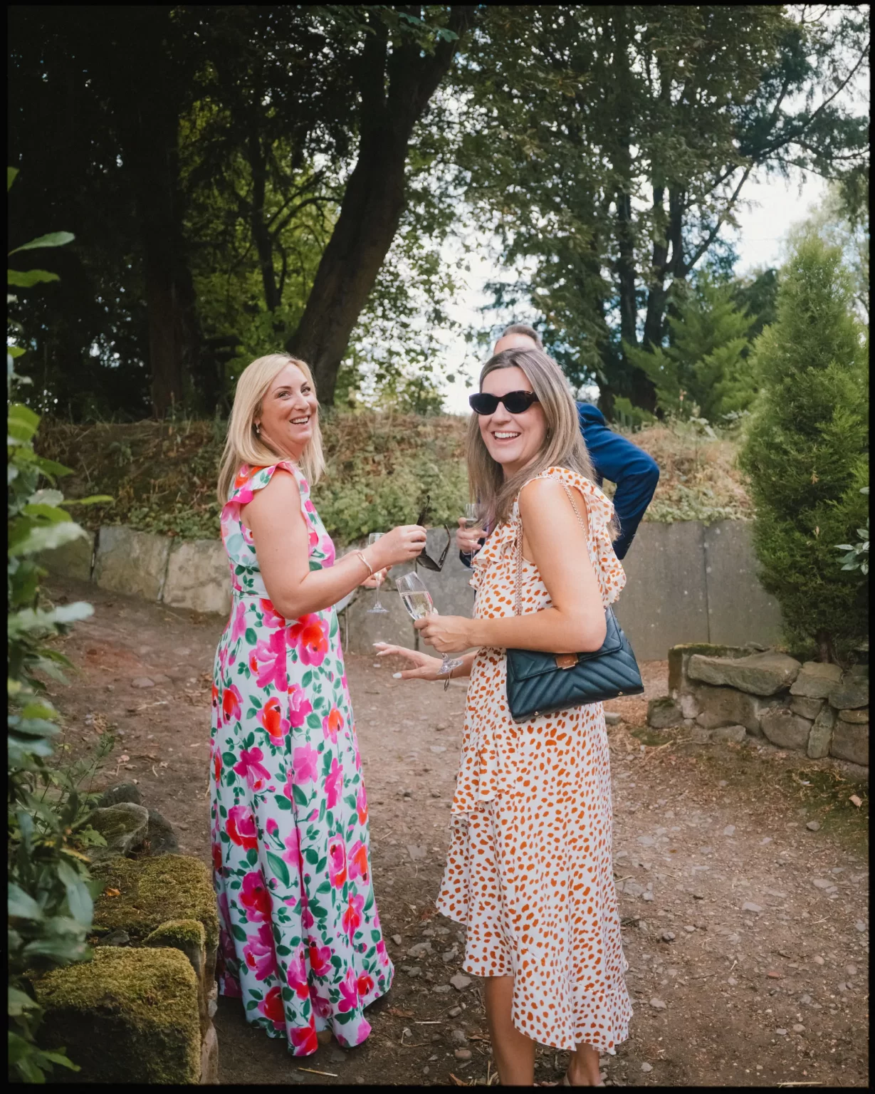 Two women in colorful dresses, one floral and one polka-dotted, stand on a garden path smiling at the camera. They are holding drinks, and tall trees fill the background.
