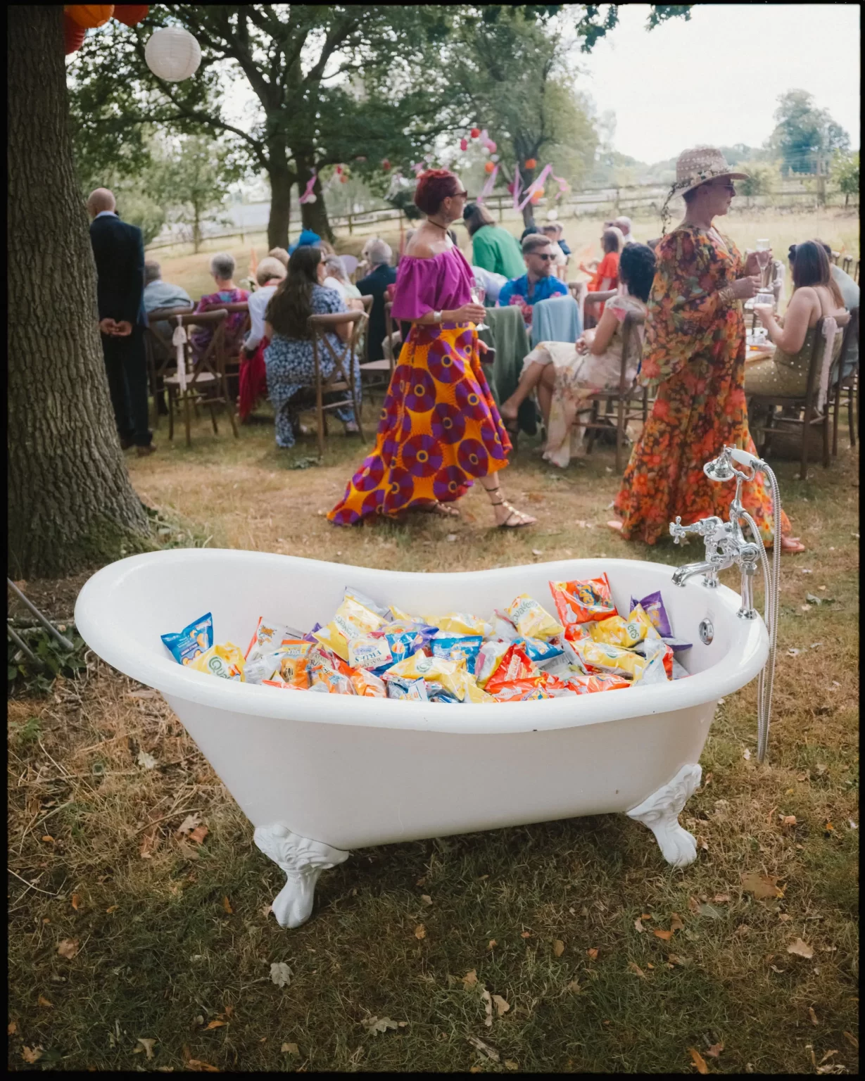 A vintage white clawfoot bathtub filled with bags of chips sits outdoors on grass, while people in colorful attire gather and sit on chairs in the background, suggesting a festive outdoor event or party at guys cliffe house, warwick
