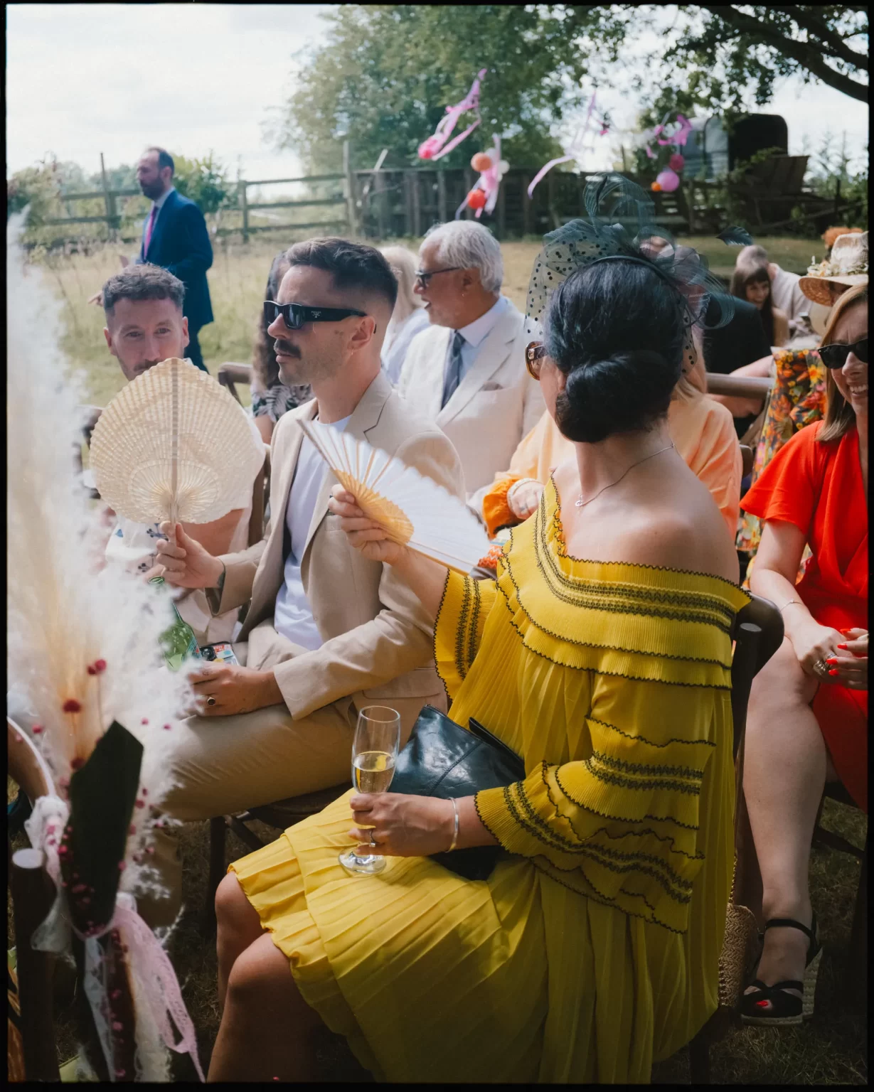 Guests at an outdoor event sit on chairs in a grassy area. A woman in a yellow dress holds a fan and a drink, while a man beside her wears a tan suit and sunglasses. Others are dressed in bright colors and summer attire.