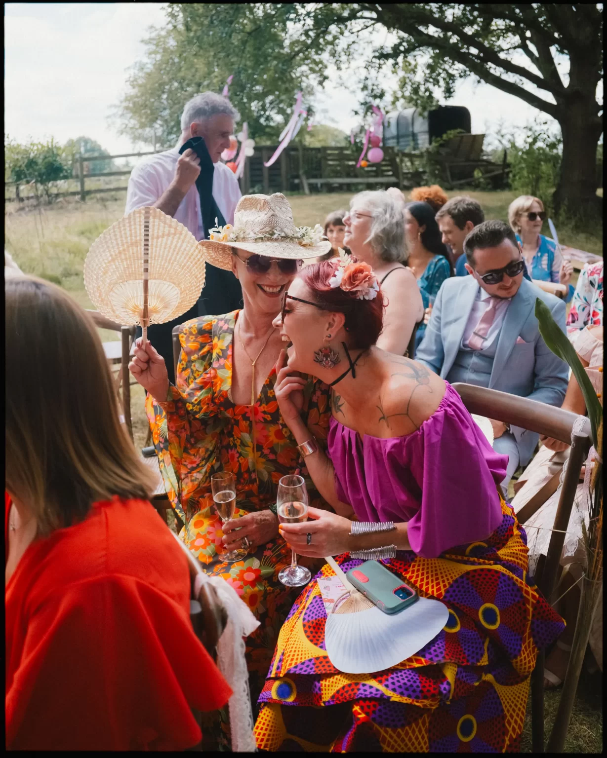 A group of people at an outdoor event sit on wooden benches, dressed in colorful, festive clothing. Two women in the foreground laugh together, holding drinks and fans, while others chat in the background under trees at guys cliffe house, warwick