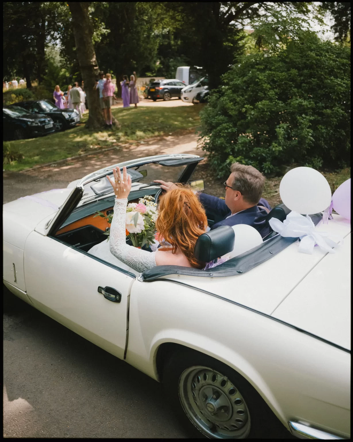 A couple in wedding attire rides in a white convertible decorated with white balloons and flowers; the bride waves as they drive away, with wedding guests in the background.
