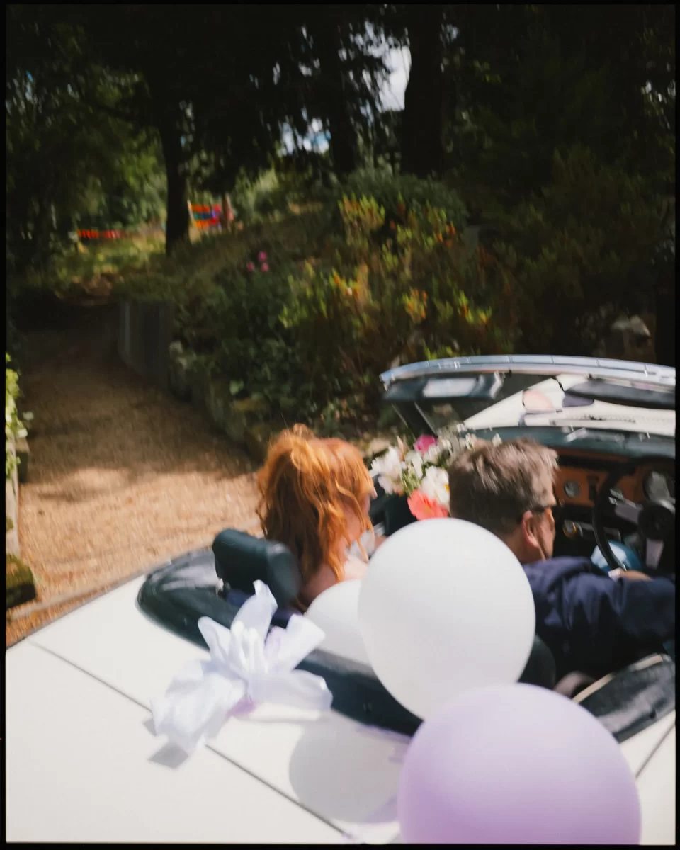 A couple sits in a white convertible decorated with white and purple balloons, driving down a gravel path surrounded by greenery and flowers, suggesting a celebratory or wedding event.