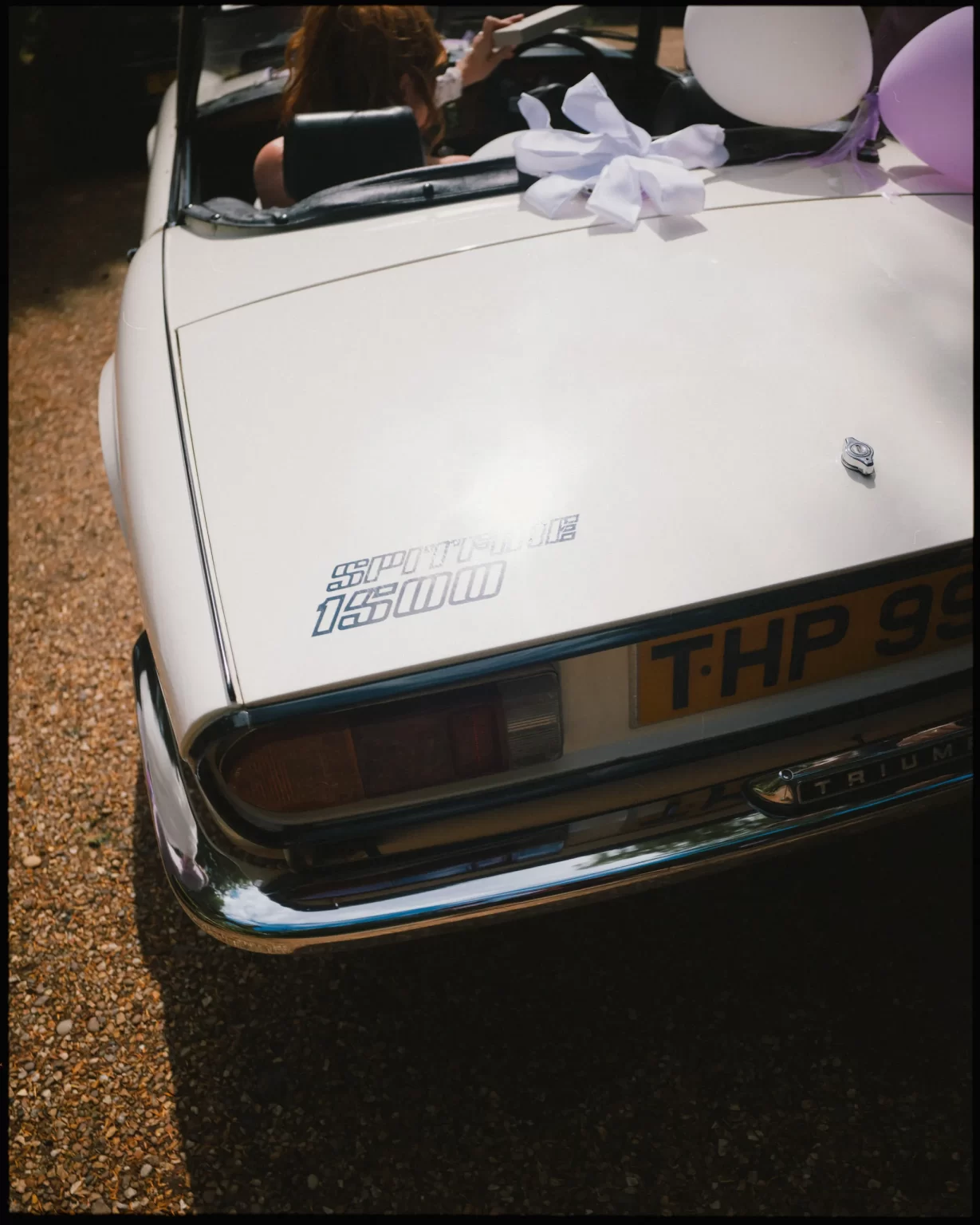A white vintage Triumph convertible decorated with balloons and a white ribbon, parked on a gravel surface. The rear badge reads Sport Six 1500. A person is partially visible in the drivers seat.