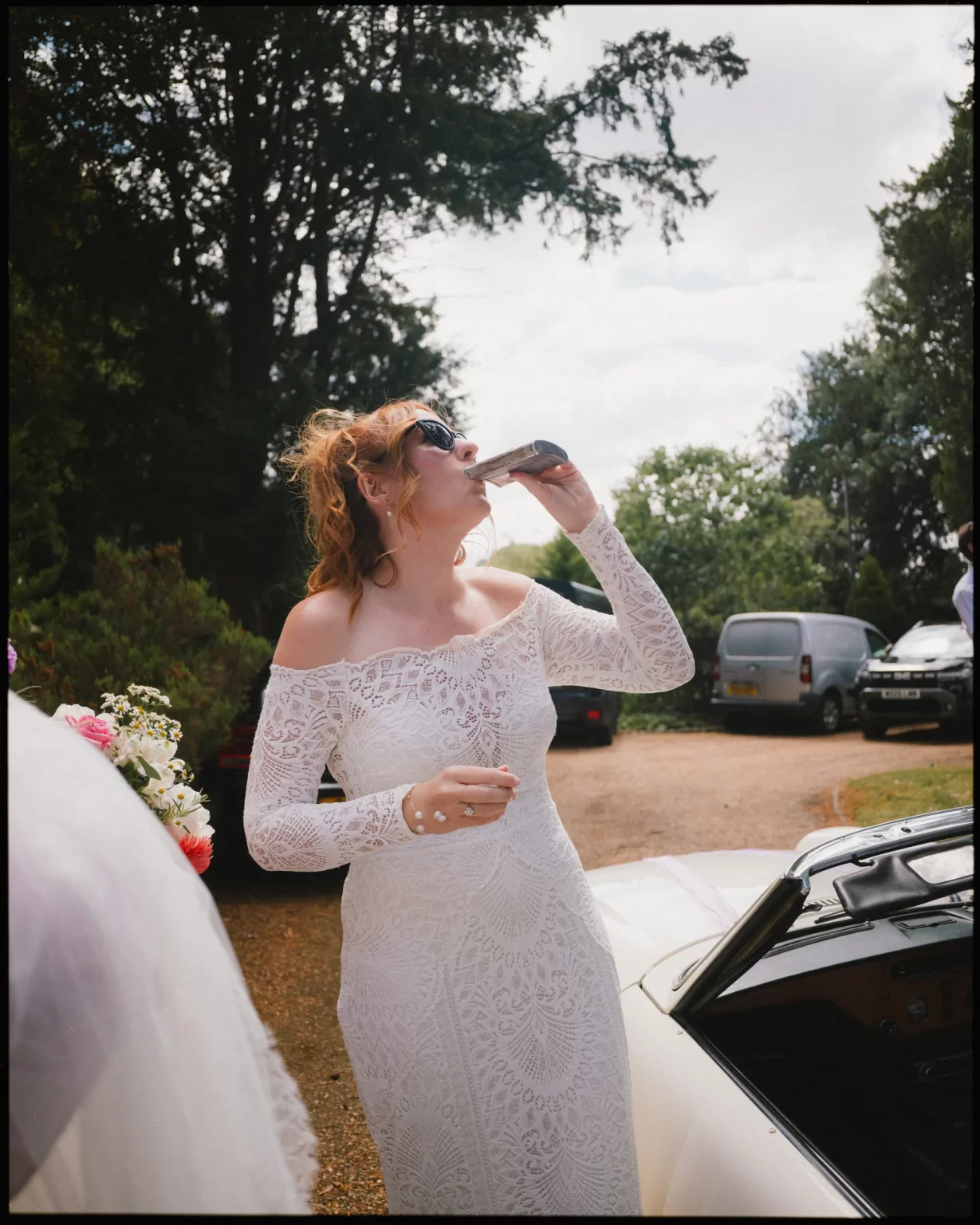 A woman in a white lace wedding dress and sunglasses stands outdoors beside a car, drinking from a metal flask. Trees and parked cars are visible in the background at guys cliffe house, warwick