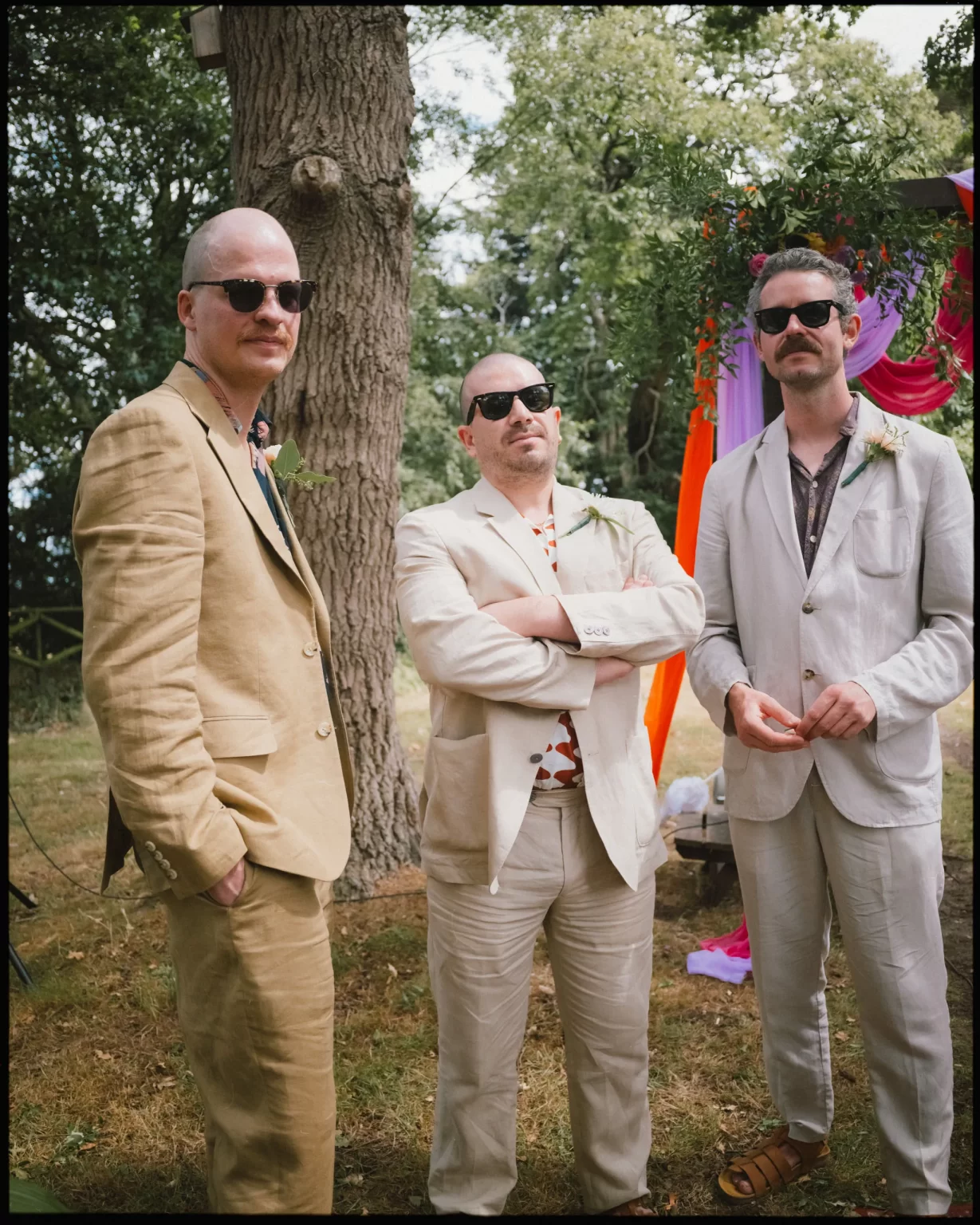 Three men in light-colored suits and sunglasses stand outdoors by a tree. They have mustaches and are at a colorful event with draped fabrics and greenery in the background at guys cliffe house, warwick