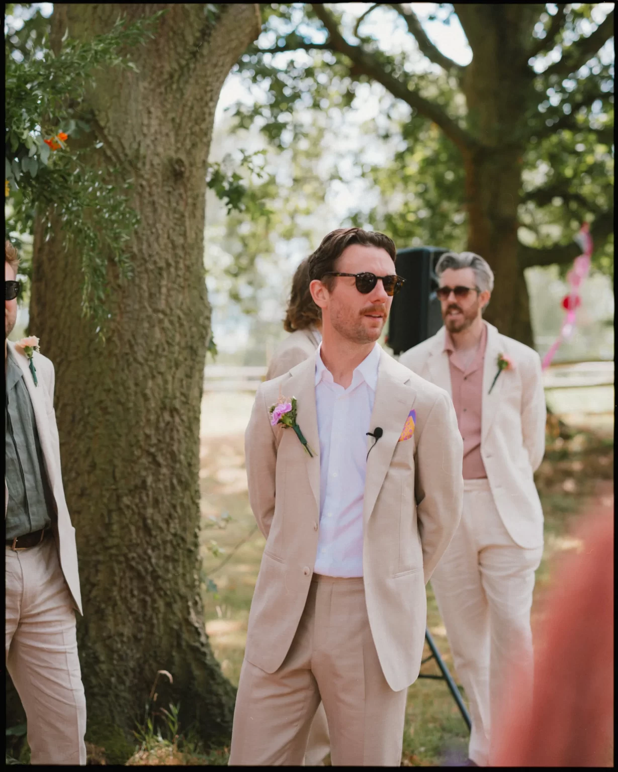 A man in a light beige suit with a boutonniere and sunglasses stands outdoors near trees, with two similarly dressed men in the background on a sunny day at guys cliffe house, warwick