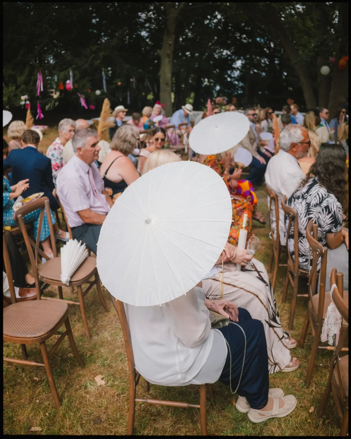 A group of people sit outdoors on wooden chairs, many holding or shading themselves with white parasols. The scene is festive, with trees and colorful decorations in the background at guys cliffe house, warwick