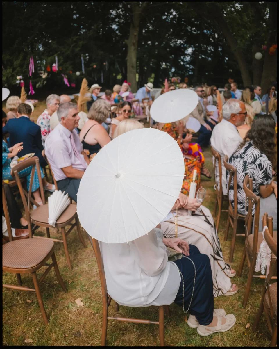 A group of people sit outdoors on wooden chairs, many holding or shading themselves with white parasols. The scene is festive, with trees and colorful decorations in the background at guys cliffe house, warwick