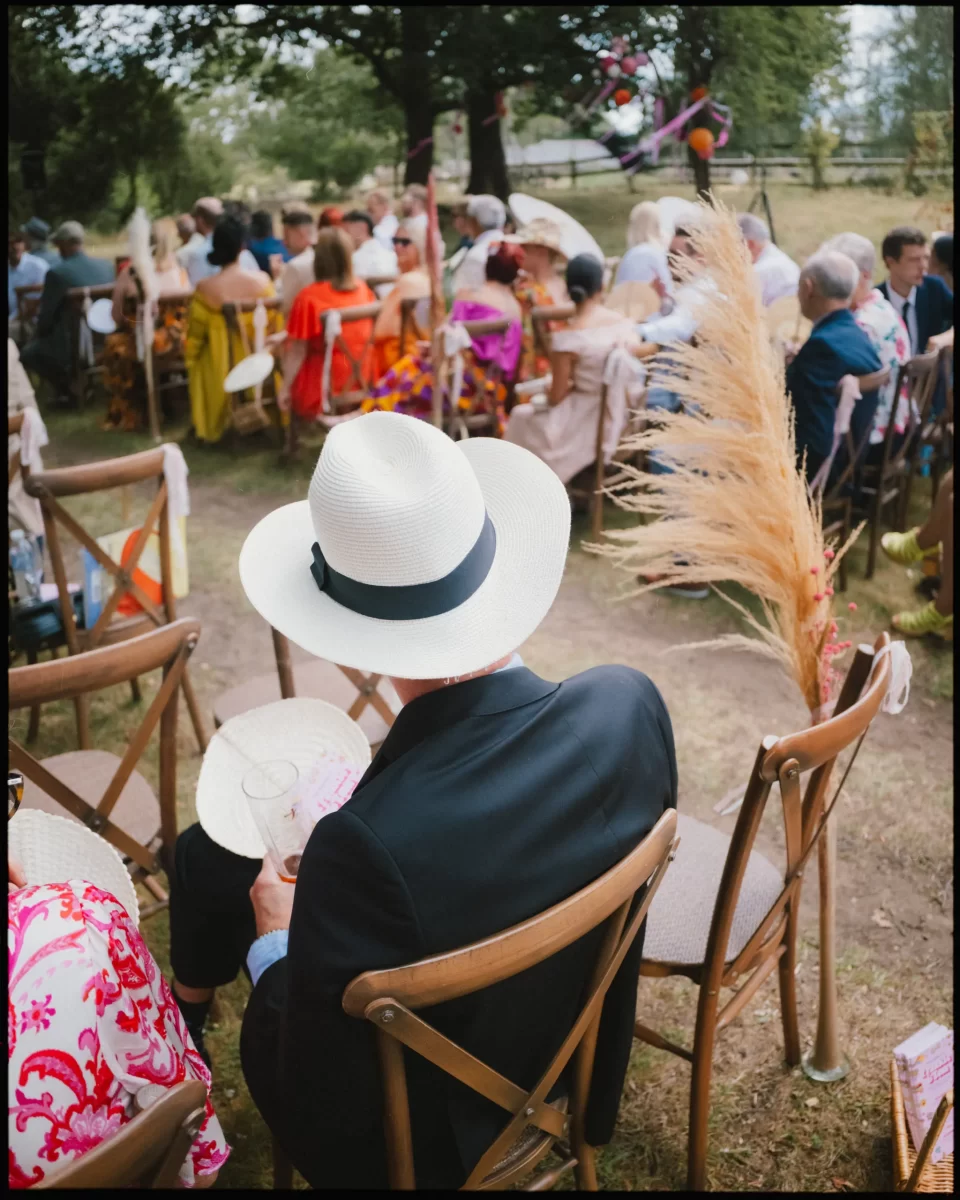 A person in a white hat and dark jacket sits on a wooden chair at an outdoor event, surrounded by people wearing colorful clothing. Dried pampas grass decorates the aisle. Trees and greenery are visible in the background at guys cliffe house, warwick