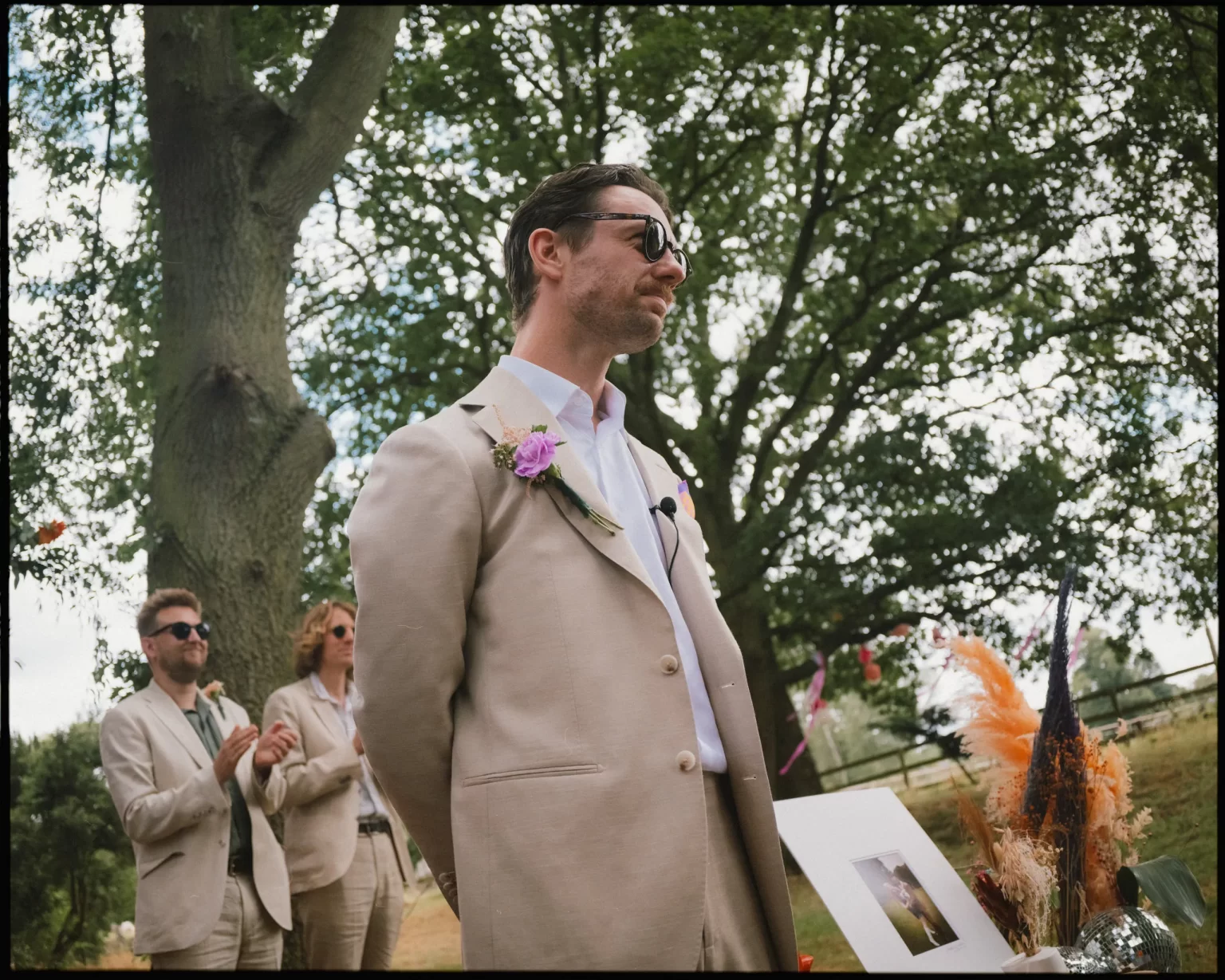 A man in a beige suit with a purple boutonniere stands outdoors by a table with flowers and a photo frame. Two people in similar attire stand and clap behind him; leafy trees fill the background at guys cliffe house, warwick