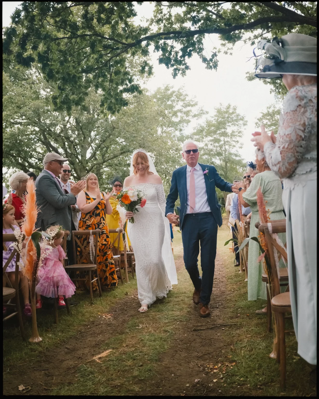 A bride in a white dress holding a bouquet walks down an outdoor aisle with an older man in a blue suit and sunglasses, surrounded by applauding guests under trees at guys cliffe house, warwick
