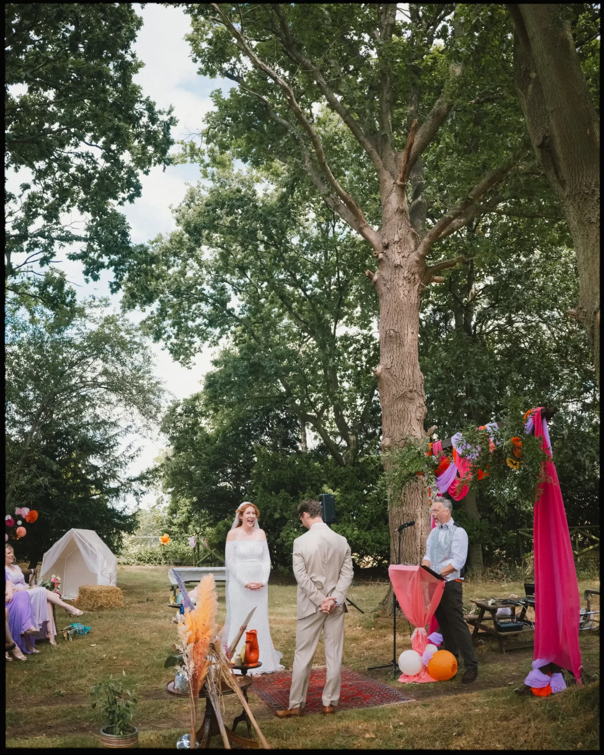 A bride and groom stand together outdoors under large trees during a wedding ceremony, with a decorated archway, colorful ribbons, and balloons. Guests sit nearby, and a person officiates beside a microphone stand at guys cliffe house, warwick