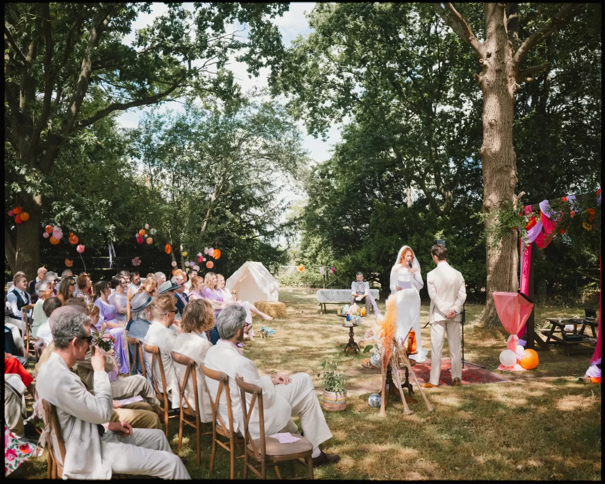 An outdoor wedding ceremony with guests seated on wooden chairs, facing a couple standing under a decorated arch. Colorful decorations and trees surround the sunny, festive scene at guys cliffe house, warwick