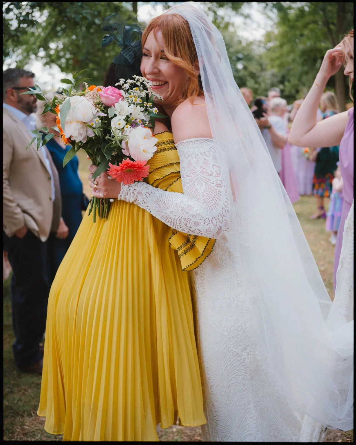 A smiling bride in a lace wedding dress and veil hugs a guest in a bright yellow dress while holding a bouquet of flowers at an outdoor celebration. Other guests are visible in the background at guys cliffe house, warwick