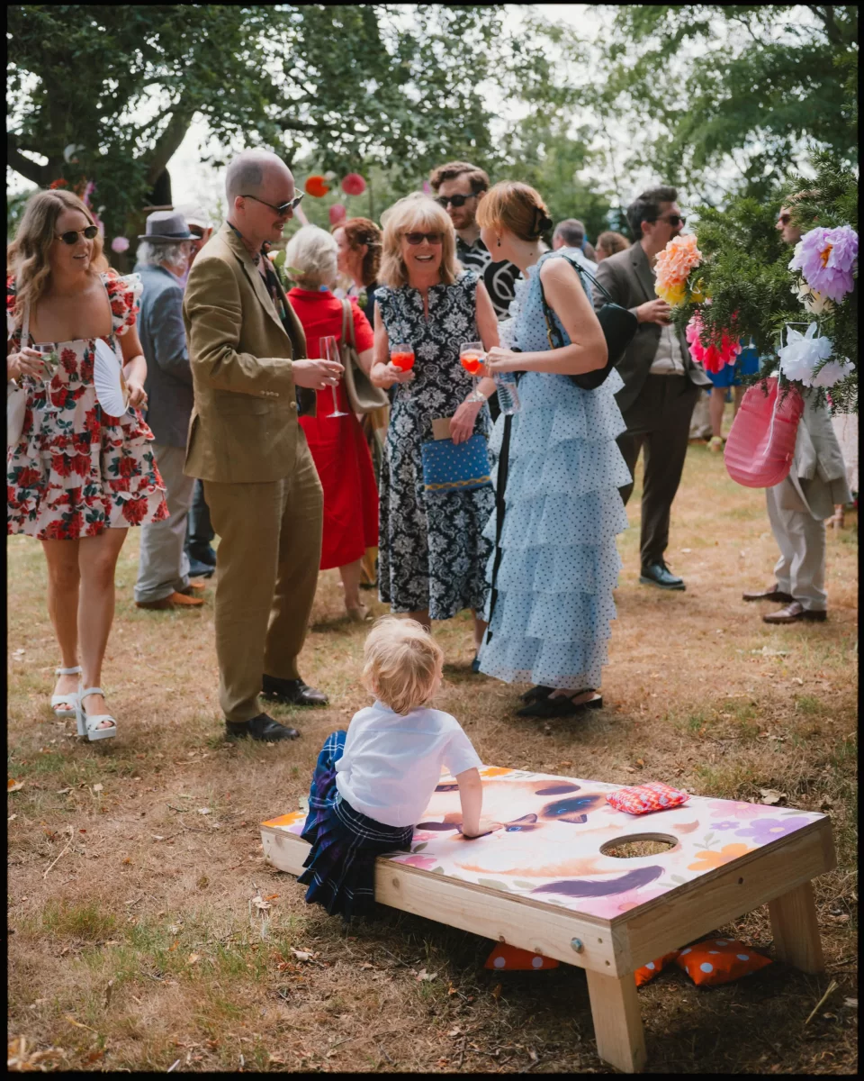 A toddler plays cornhole on the grass while adults dressed in colorful, summery outfits and holding drinks stand nearby, talking and smiling at an outdoor gathering under trees at guys cliffe house, warwick