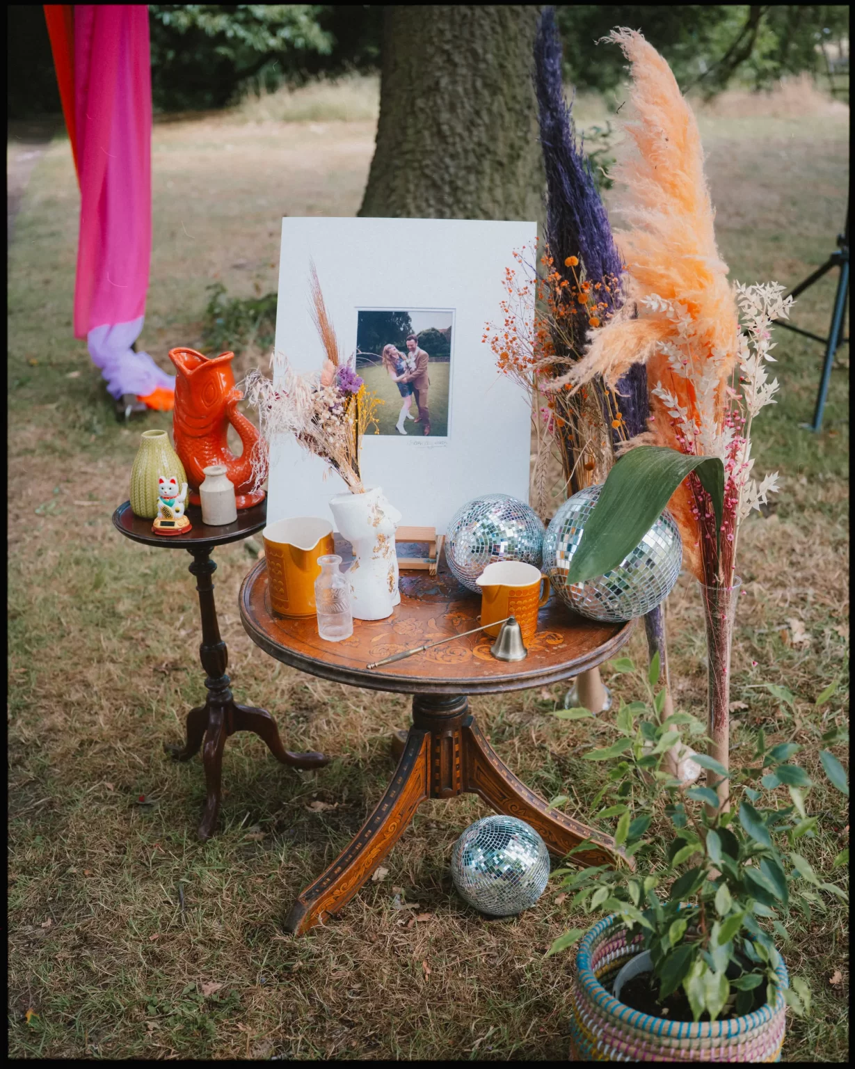 A decorative outdoor table setup with a framed photo, disco balls, colorful vases, dried pampas grass, candles, a ceramic fox, and a potted plant, arranged under a tree on grass.