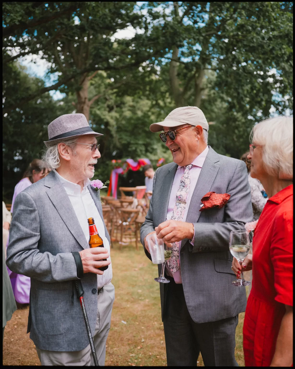 Three older adults dressed in suits and bright clothing talk and laugh outdoors, each holding a drink. Trees and colorful decorations appear in the background, suggesting a festive event or garden party at guys cliffe house, warwick