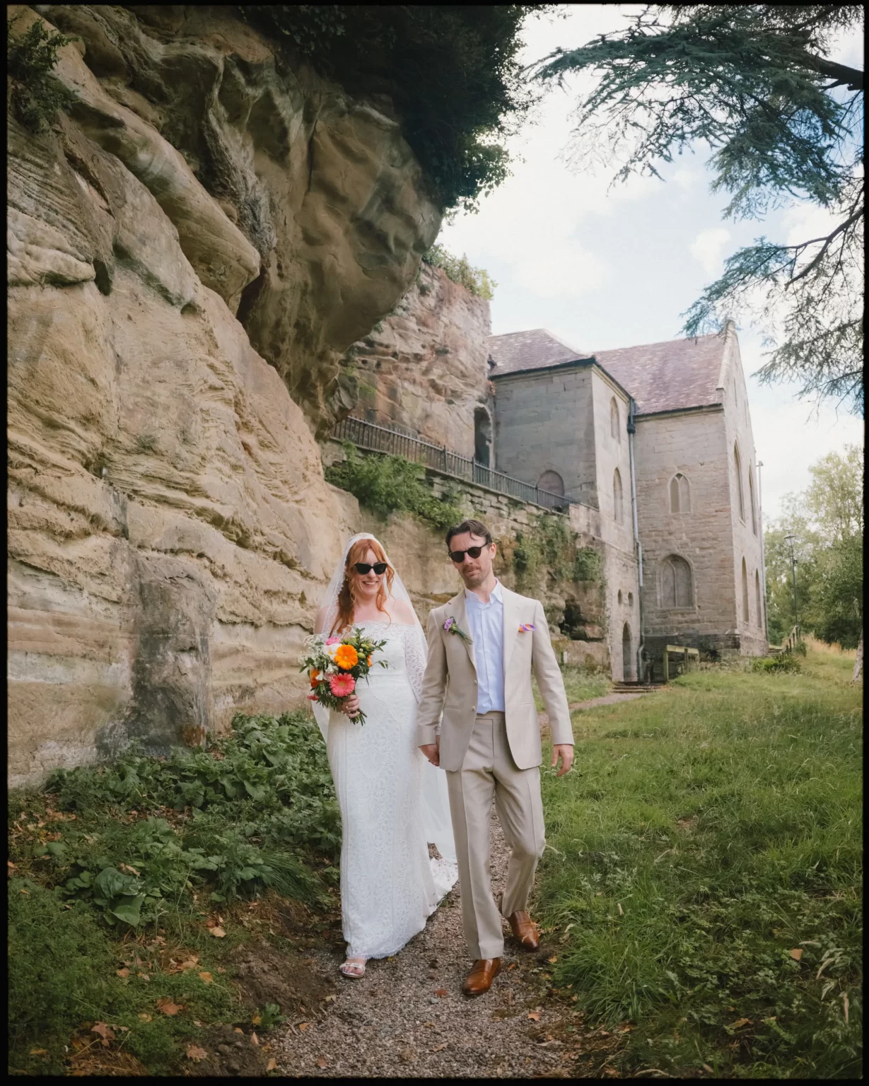 A bride in a white dress holding a bouquet walks beside a groom in a tan suit along a path by stone cliffs and the historic Guys Cliffe House near Warwick, surrounded by lush greenery at guys cliffe house, warwick