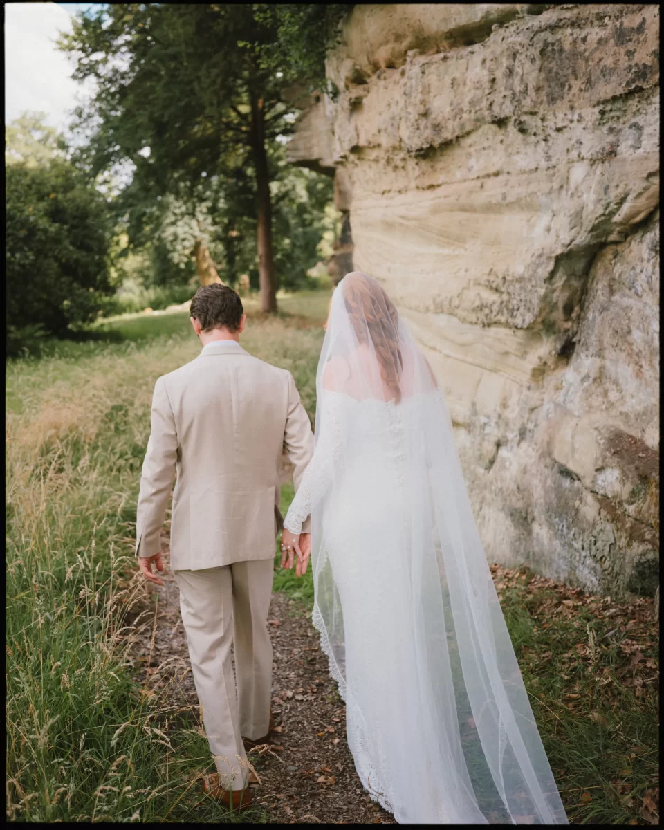 A bride in a long white dress and veil walks hand in hand with a groom in a tan suit along a grassy path beside a rocky cliff, surrounded by trees and greenery at guys cliffe house, warwick