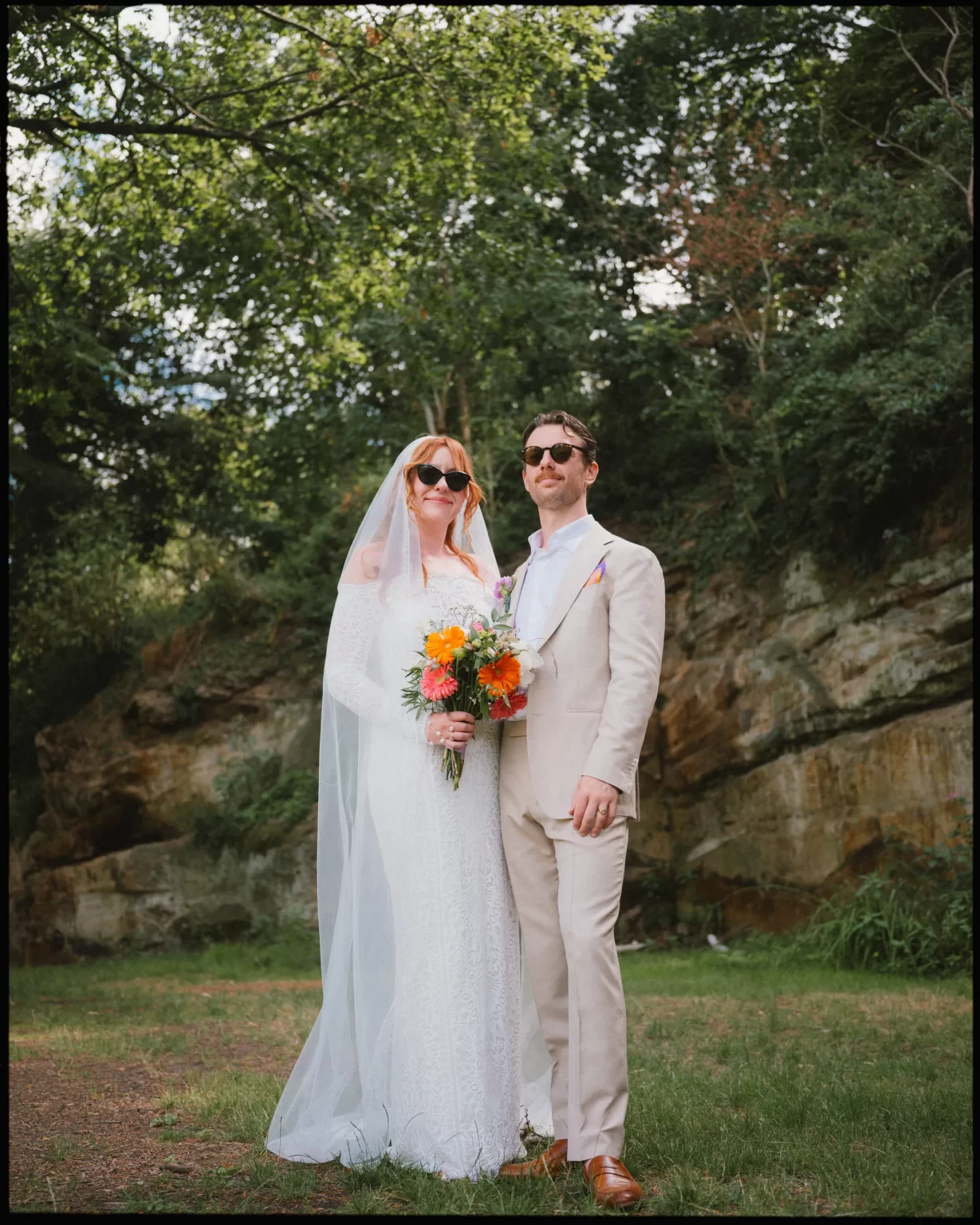 A bride and groom stand outdoors, both wearing sunglasses. The bride holds a colorful bouquet and wears a white dress with a veil. The groom is in a light suit. Lush green trees and a rocky background are behind them at guys cliffe house, warwick