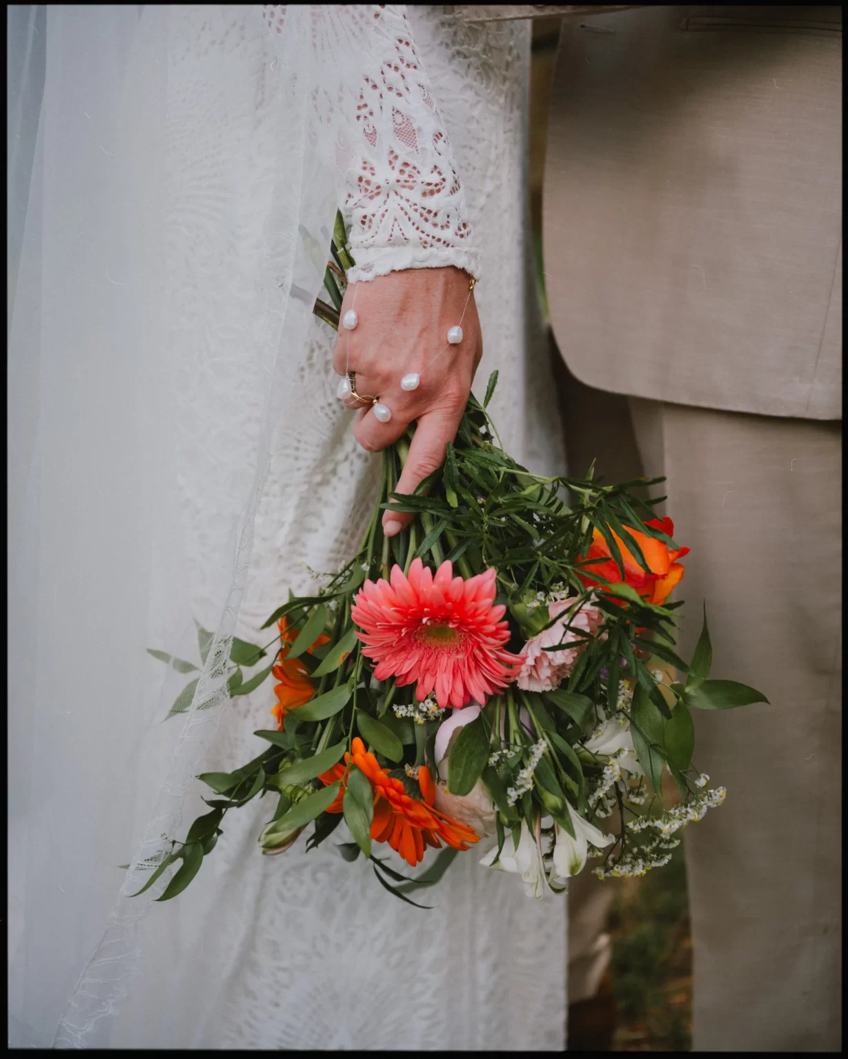A close-up of a bride’s hand holding a bouquet of pink and orange flowers, with greenery. She is wearing a lace wedding dress and pearl bracelet, standing beside someone in a beige suit.